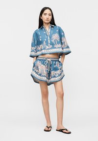 Γυναίκα που φοράει μπλε crop top και ταιριαστά σορτς με λευκό κορδόνι και ροζ και λευκά τοπία ως σχέδια, στέκεται με τα χέρια στις τσέπες.