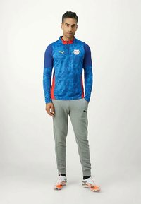 Άνδρας που στέκεται φορώντας μπλε και κόκκινη μακρυμάνικη αθλητική μπλούζα Puma, γκρι παντελόνι Puma και λευκά με πορτοκαλί παπούτσια. 