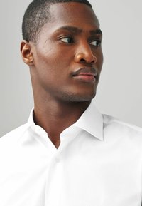 Chemise blanche à col boutonné, texture lisse et design ajusté, présentée sur un mannequin avec les cheveux courts et des détails faciaux.
