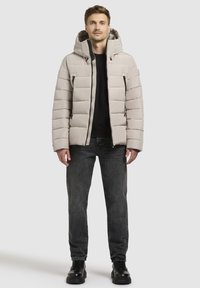 Svetlo siva pufasta jakna s kapuco, zadrgo spredaj in stranskimi žepi. Ujemajo se z črnimi kavbojkami in črnimi čevlji. Gladka tekstura in prešita zasnova.