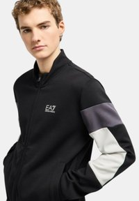 Giacca nera con zip caratterizzata da un colletto alto, tessuto testurizzato e maniche a blocchi di colore grigio e bianco. Logo EA7 sul petto.