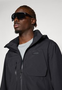 Black waterproof jacket with a zip front, high collar, and visible branding. Features a large front pocket and is paired with dark sunglasses.