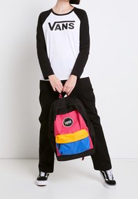 Sort og hvid langærmet skjorte med "VANS" logo; farverig rygsæk med pink, gul og blå sektioner og cirkulær logo. Sorte bukser og sko.
