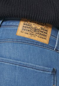 Blå jeans i lätt blå denim. Etiketten är av tanfärgad läder och har ett svart tryckt arkitektoniskt mönster samt textmarkeringar.