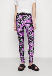 Leggings nere con un vivace motivo floreale viola e dettagli verdi. La vita è nera con testo bianco "crás" e il tessuto sembra lucido.