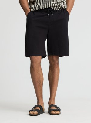 Mężczyzna ubrany w czarne szorty ze sznurkiem, czarne sandały i czarno-biały pasiasty bezrękawnik, stojący z rękami w kieszeniach.