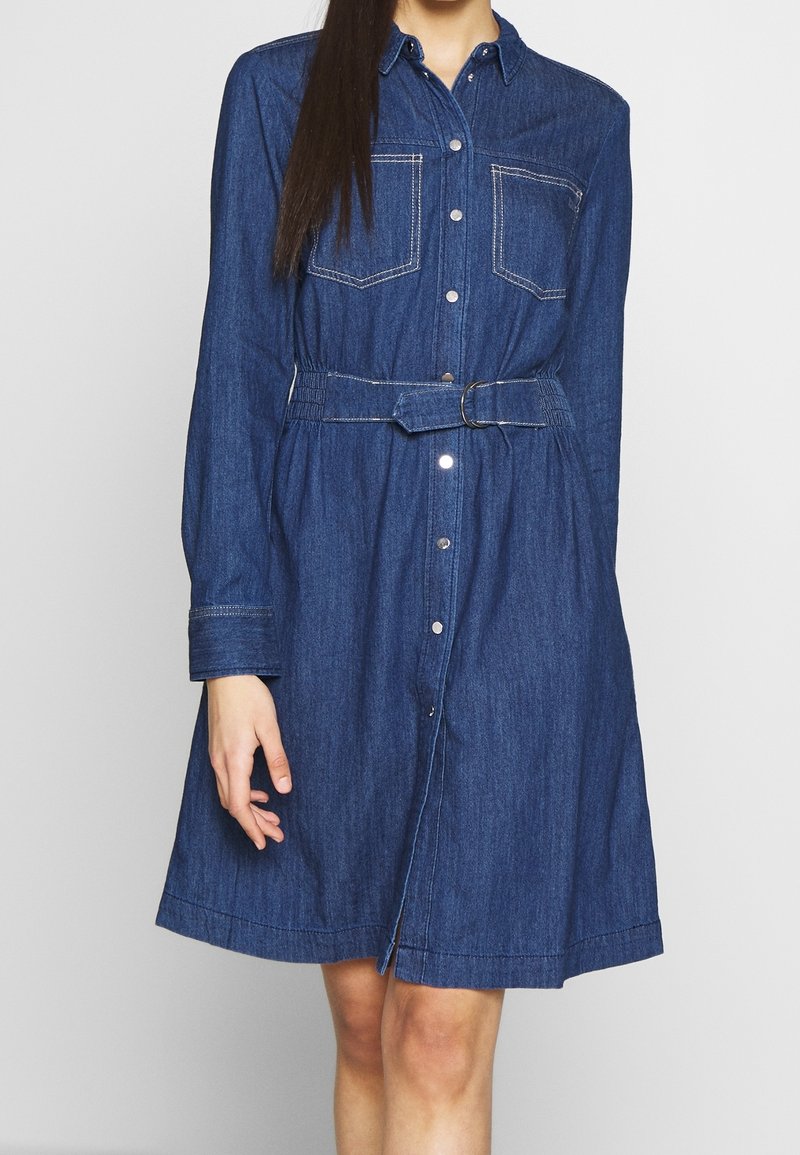Denimini mekko, jossa on vyöllinen vyötärö, pitkät hihat, napitus edessä ja kaksi rintataskua. Tumma sininen kangas kontrastivärisine ompeleineen.