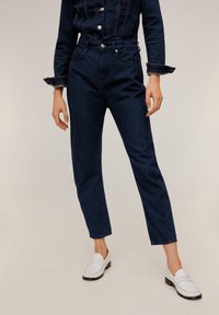 Jeans de mezclilla azul oscuro con cintura alta, corte ajustado y diseño clásico de cinco bolsillos, combinados con mocasines blancos. Textura suave, detalles mínimos.