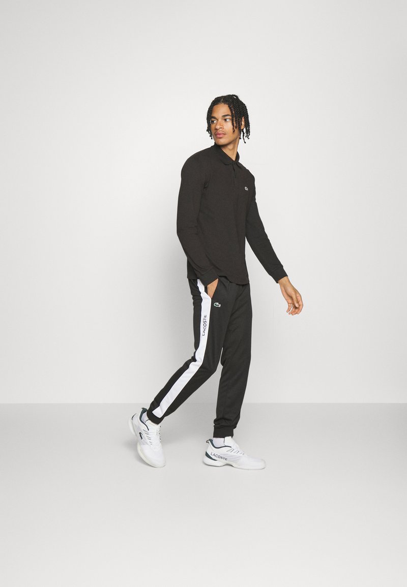 Jeune homme portant un polo noir à manches longues, un pantalon de jogging noir avec des rayures blanches sur les côtés, des baskets Lacoste blanches, marchant devant un fond gris clair.