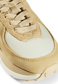 Gros plan sur le bout d’une basket beige en daim et mesh avec des lacets plats et une semelle en caoutchouc blanche visible.