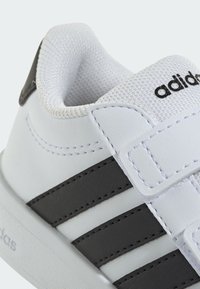 Bijela sportska cipela od kože s tri crne pruge, podstavljenim okovratnikom i zatvaračem na čičak. Teksturirano unutarnje postavljanje i detalji ojačanih šavova.