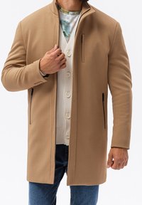 Tan kappa med en slim fit, slät textur, hög krage, dragkedjefickor på sidan och en knäppt cardigan under; bärs över blå jeans.