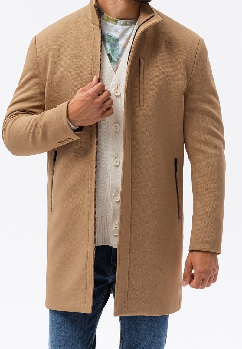 Tan kappa med en slim fit, slät textur, hög krage, dragkedjefickor på sidan och en knäppt cardigan under; bärs över blå jeans.