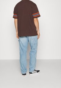 Camiseta marrón de gran tamaño con rayas azules y naranjas en las mangas, combinada con vaqueros anchos de color azul claro y zapatillas de cuadros en blanco y negro.