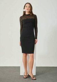 Robe noire avec de longues manches en dentelle, corsage ajusté et ourlet à mi-genoux. Texture lisse, design minimaliste, placée sur un fond neutre.