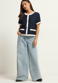 Mujer que lleva un cárdigan de manga corta azul marino con ribete blanco y botones dorados, vaqueros de pierna ancha azul claro y zapatos negros, de pie con una mano en el bolsillo.