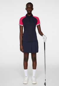 Marinblå golfpolo med rosa ärmar, kragdesign och knappslå. Kombinerad med en plisserad marinblå kjol. Modellen håller en golfklubba och bär vita strumpor och skor.