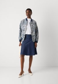 Cvetna bomber jakna iz modrega in belega blaga, bela majica, razširjena jeans krila in bež pete mule, drži torbico svetle barve.