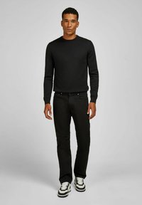 Jeune homme debout, portant une chemise noire à manches longues, un pantalon noir et des sneakers noir et blanc sur un fond gris uni.