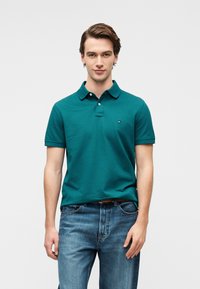 Τιρκουάζ πόλο μπλούζα από βαμβάκι με πατιλέτα δύο κουμπιών και κοντά μανίκια. Διαθέτει μικρό λογότυπο στο στήθος. Συνδυάζεται με μπλε τζιν παντελόνια.