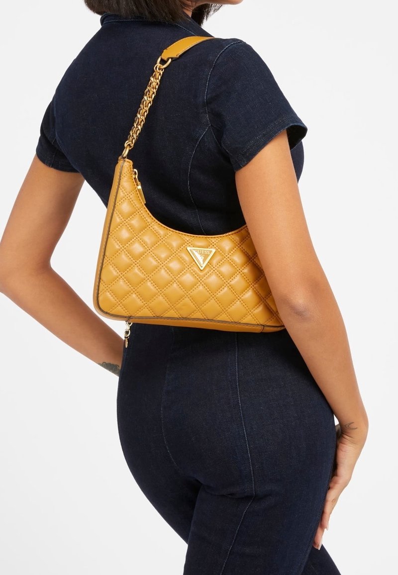Femme portant une tenue en denim foncé, portant un sac à bandoulière en cuir matelassé couleur camel avec une chaîne dorée et un logo triangulaire.
