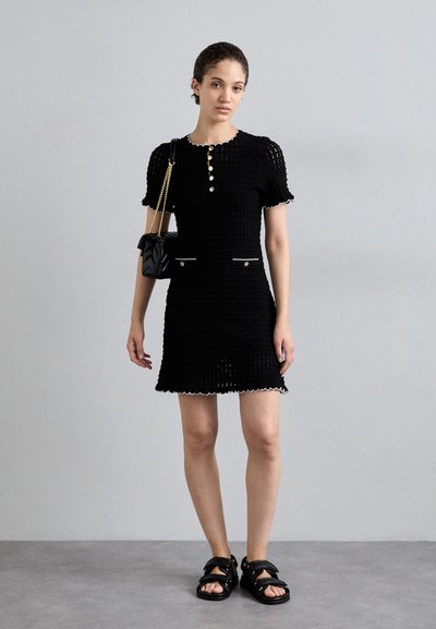 Schwarzes Strickkleid mit kurzen Ärmeln, gewelltem Saum, Knopfdetails und weißen Akzenten. Getragen mit einer schwarzen Mini-Tasche und flachen Sandalen.