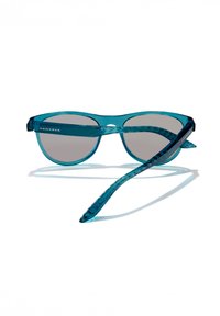 Gafas de sol color aqua con montura transparente, lentes grises y patillas texturizadas. Logo "HAWKERS" visible en la parte frontal.