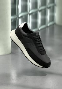 Μαύρο αθλητικό παπούτσι με επάνω μέρος από υφή δέρματος, αντίθετη λευκή σόλα και επίπεδα κορδόνια. Διαθέτει ομαλό σχεδιασμό και στρογγυλεμένη μύτη.