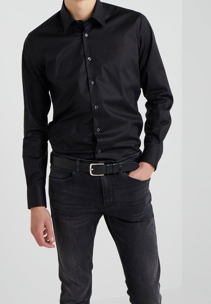 Homme portant une chemise noire boutonnée rentrée dans un jean gris foncé avec une ceinture noire, une main dans la poche, debout devant un fond uni.