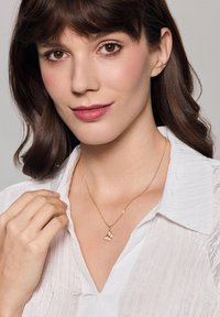 Złoty naszyjnik z zawieszką w kształcie motyla, delikatny łańcuszek, noszony na białej, teksturowanej bluzce. Prosty design, polerowane wykończenie, subtelne akcenty.