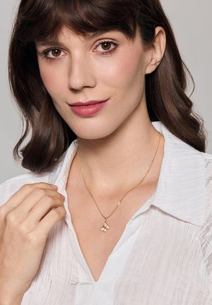 Złoty naszyjnik z zawieszką w kształcie motyla, delikatny łańcuszek, noszony na białej, teksturowanej bluzce. Prosty design, polerowane wykończenie, subtelne akcenty.