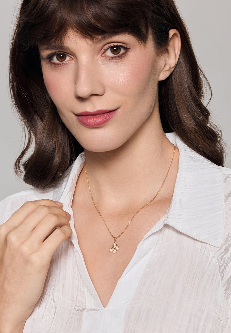 Złoty naszyjnik z zawieszką w kształcie motyla, delikatny łańcuszek, noszony na białej, teksturowanej bluzce. Prosty design, polerowane wykończenie, subtelne akcenty.