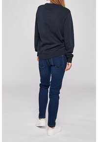 Sweat-shirt bleu marine avec poignets et ourlet côtelés, associé à un jean bleu foncé. Modèle portant des baskets blanches, vu de dos.