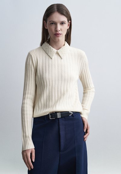 Femme portant un pull crème côtelé avec un col orné de perles et un pantalon plissé bleu marine ceinturé de noir, debout devant un fond clair.