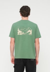 Πράσινο βαμβακερό μπλουζάκι με γραφική εκτύπωση βουνών, δέντρων και ήλιου στην πλάτη. Λαιμόκοψη crew, κοντά μανίκια, άνετη εφαρμογή.