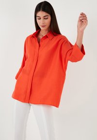 Bright orange, oversized linen shirt with a pointed collar, button closure, and three-quarter sleeves, paired with white slim-fit pants.