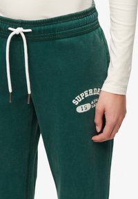 Grønne sweatsbukser lavet af blødt stof med elastisk talje, hvid snøre og et logo printet i hvid nær hoften.