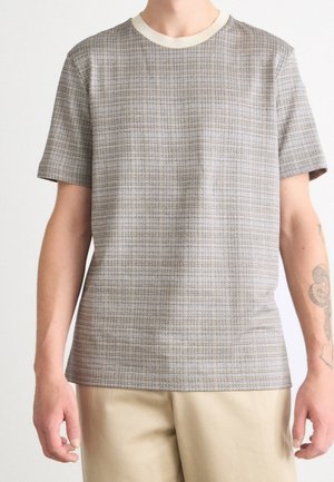 Mann, der ein kurzärmeliges beige-graues T-Shirt mit cremefarbenem Kragen und hellbeigen Hosen trägt und ein Tattoo am rechten Arm zeigt.