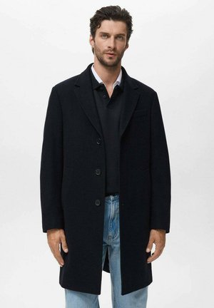 Abrigo de lana azul marino con cuello de pico, diseño de un solo botón y dos bolsillos delanteros. Llevado sobre un polo negro y jeans azules.