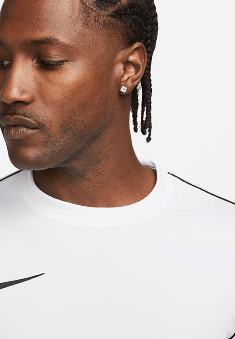 Bílý atletický dres Nike s kulatým výstřihem a černými akcentovými pruhy, doplněný náušnicemi a krátkými, zapletenými vlasy.