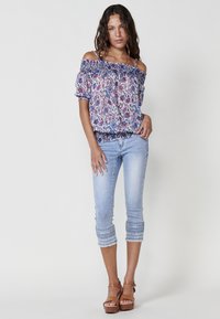 Blusa floreale off-the-shoulder in tessuto leggero, multicolore con dettagli blu; abbinata a jeans cropped azzurri con orli ricamati.