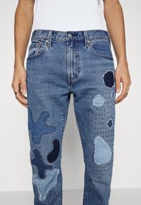 Jeans i denim med lappar i olika blå nyanser, patchade designs och texturerade detaljer på framsidan. Klassisk femficksmodell.