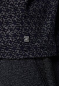 Tesătură cu model marin și design în spirală; prezintă un mic etichetă pătrată cu logo la tiv. Textura pare moale, cu o suprafață netedă.
