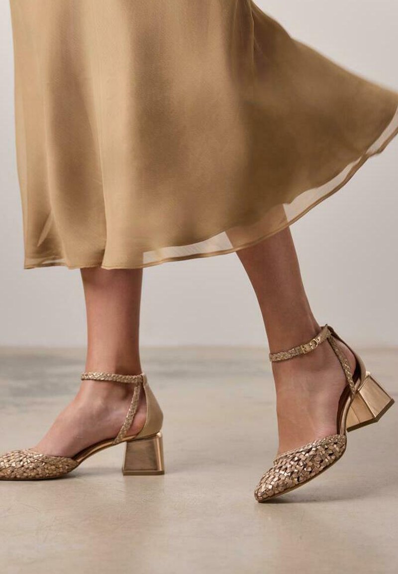 Sandales dorées à talons épais tressés avec une bride à la cheville. Une robe beige semi-transparente tombe autour des jambes.