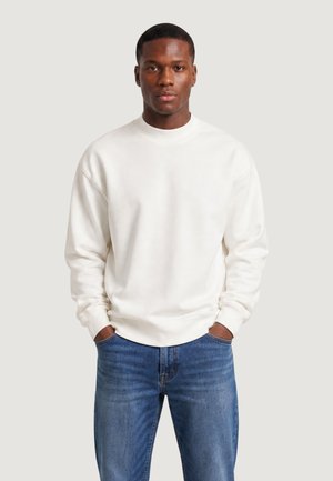 Joven con pelo corto que lleva una sudadera blanca y vaqueros azules, de pie con las manos en los bolsillos frente a un fondo sencillo.