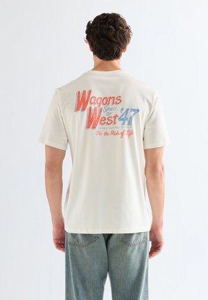 Man met kort donker haar draagt een crèmekleurig T-shirt met rode en blauwe tekst "Wagons West 47" op de achterkant, gecombineerd met lichtblauwe spijkerbroek.