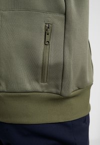 Felpa con zip verde oliva realizzata in un tessuto testurizzato, con un orlo inferiore a costine, dotata di una tasca laterale con zip per riporre gli oggetti.