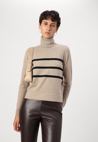 Beige garbó pulóver fekete vízszintes csíkokkal, puha anyagból készült, magasan ültetett barna bőr nadrággal és krémszínű táskával párosítva.