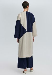 Lange, oversized jas in beige en marineblauw met een geometrisch kleurblokontwerp. Draagt over wijde marineblauwe broeken. Glad, textuurstof.