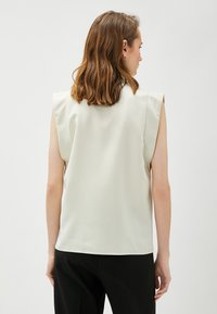 Ärmellose Bluse aus leichtem cremefarbenem Stoff, mit lockerer Passform, glatter Textur und subtilen Details an den Schultern. Kombiniert mit schwarzen Hosen.